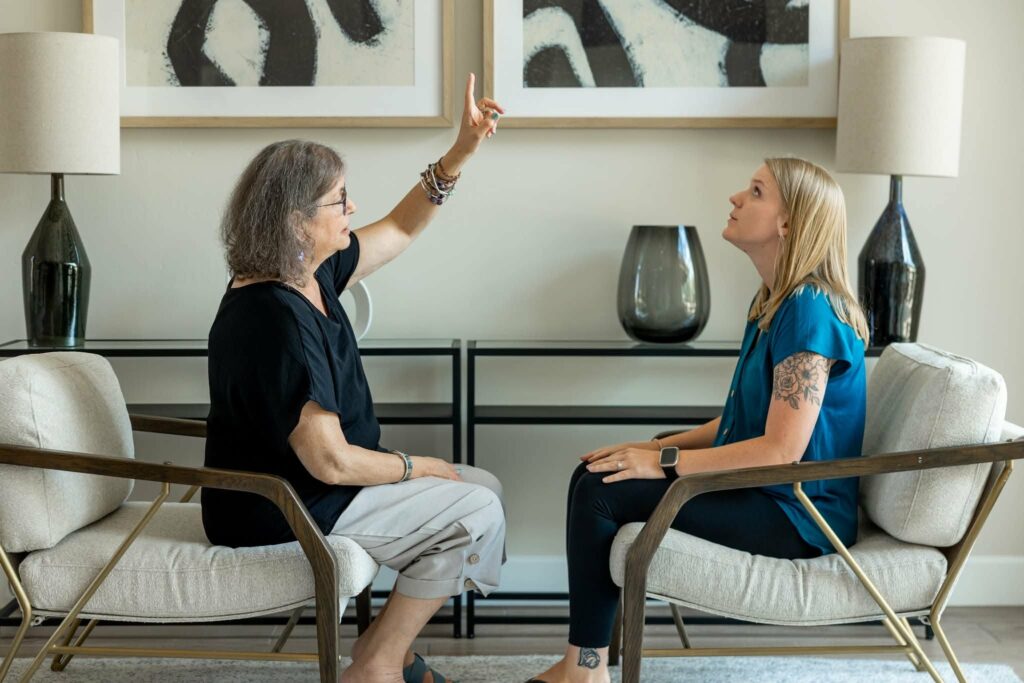 Gretel and a woman are sitting across from one another. Gretel is speaking and pointing upwards. The woman looks where Gretel is pointing.